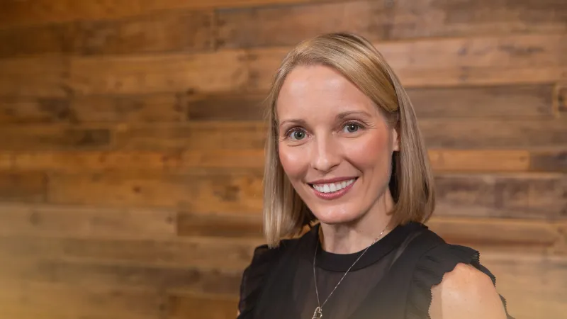 Smiling woman with blonde hair wearing a black sleeveless top and heart necklace against wooden background