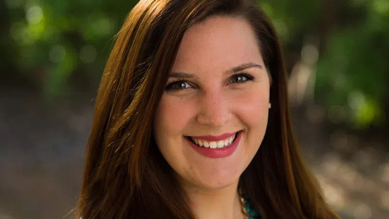 Smiling woman with long brown hair wearing a white coat and turquoise necklace outdoors in natural light