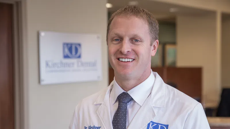 Smiling male dentist wearing white coat and tie standing in dental office with Kirchner Dental sign in background