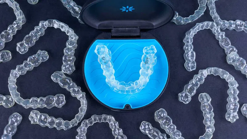 Clear dental aligners scattered around a blue case holding a pair of aligners on a dark surface.