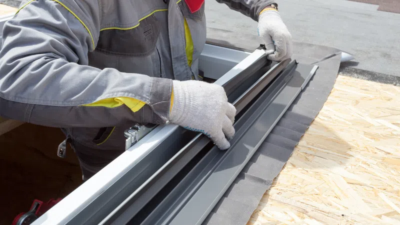 Construction worker in gloves installing window frame on plywood roof surface during building work.