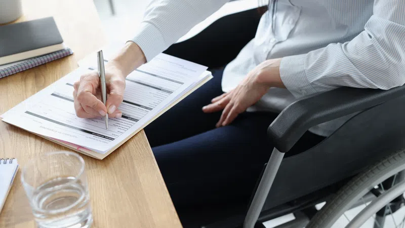 Person in wheelchair filling out a form at a wooden desk with a glass of water nearby.