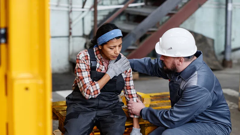 Female industrial worker sitting in pain while male coworker with helmet comforts her at factory workspace.