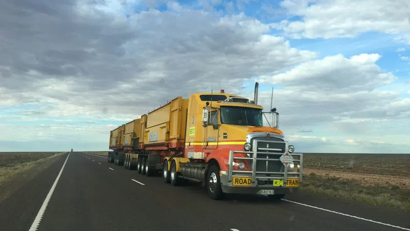 Long yellow and orange road train truck driving on empty highway under cloudy blue sky.