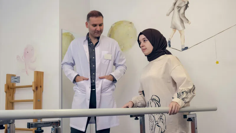 Physical therapist assists a woman in a hijab during rehabilitation exercises using parallel bars indoors.