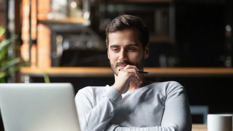 Man in white sweater thoughtfully working on laptop at cozy workspace with coffee mug and notes.