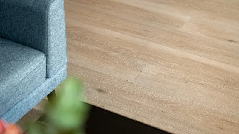 Close-up of light wood flooring beside a blue fabric sofa and blurred green plant in living room.