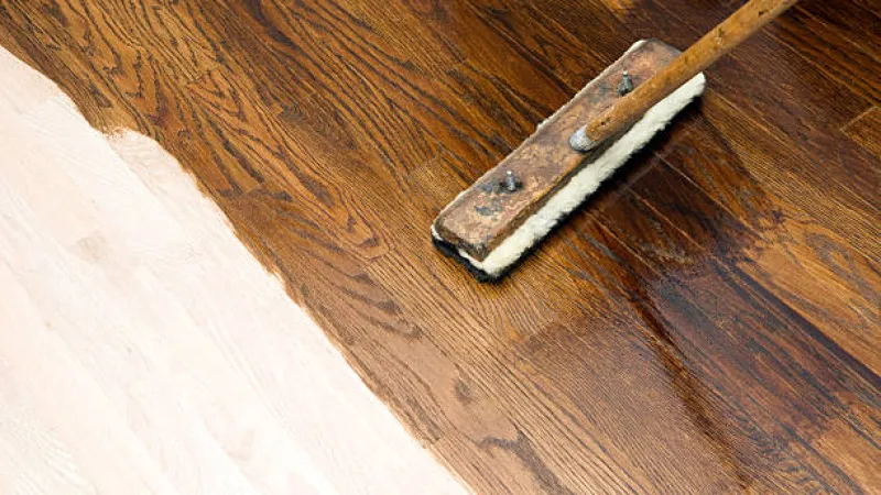 Applying dark wood stain with brush on light hardwood floor for refinishing and restoration.