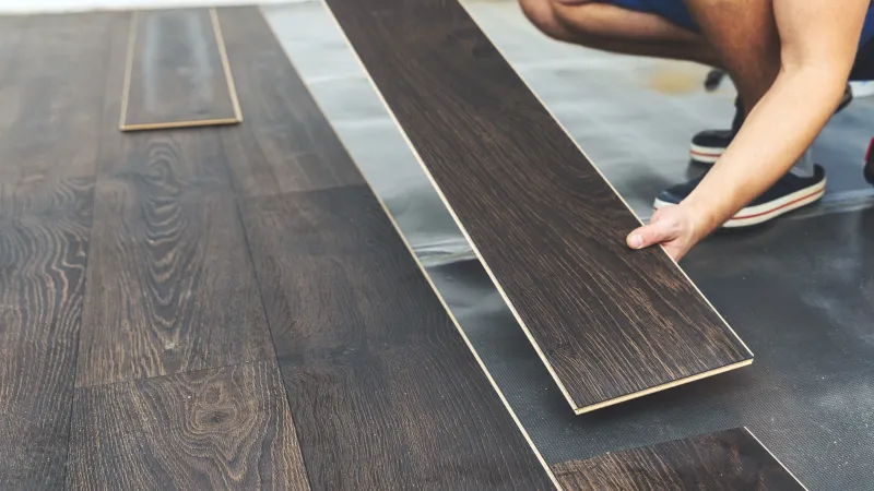Person installing dark wood laminate flooring panels on a floor with plywood underlayment visible.