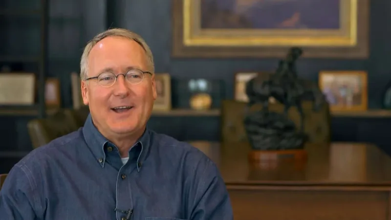 Middle-aged man wearing glasses and a blue button-up shirt smiling in a cozy room with framed art and sculpture.