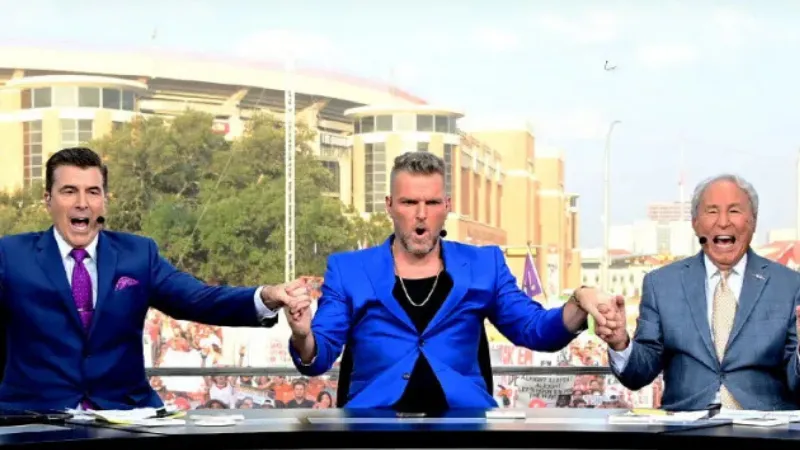 Three ESPN College GameDay hosts in suits holding hands excitedly behind the broadcast desk outdoors with stadium background.