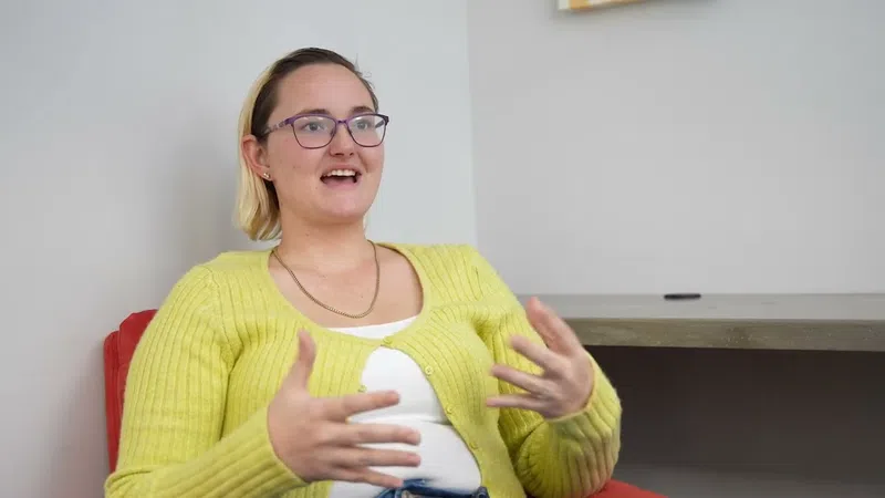 Young woman in yellow cardigan speaking animatedly while seated on a red chair indoors.