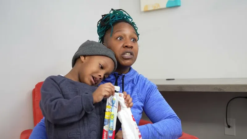 Young child with gray beanie and dark sweater leaning on woman with teal hair and blue jacket indoors