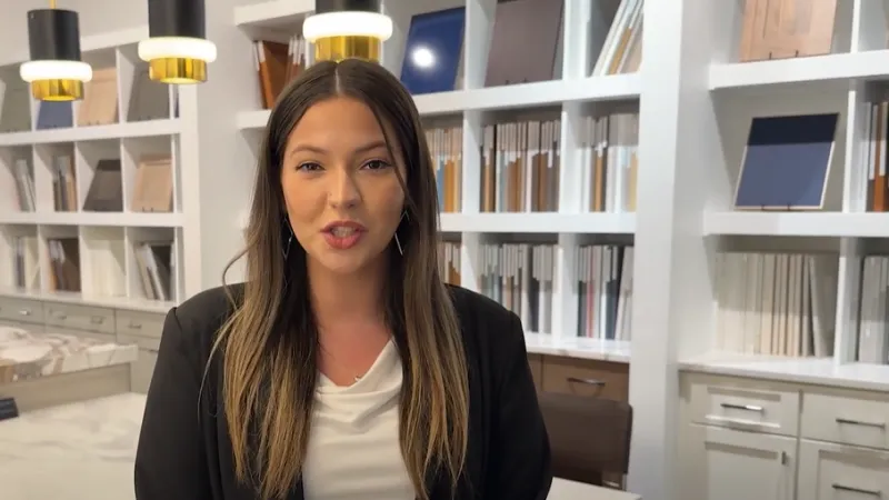 Woman in a black blazer standing in a modern showroom with marble countertops and samples on shelves behind her.