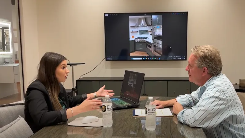 Two professionals discussing kitchen design in an office with a laptop and kitchen image on a screen behind them.