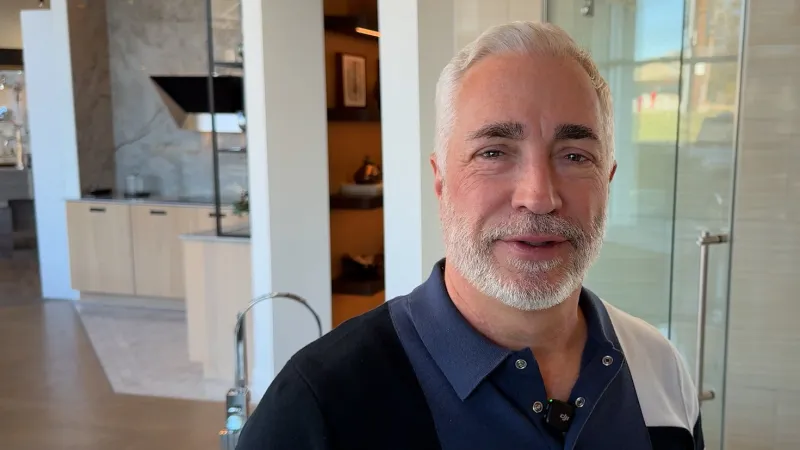 Smiling middle-aged man with gray hair and beard standing in a modern bathroom showroom with sinks and glass shower.