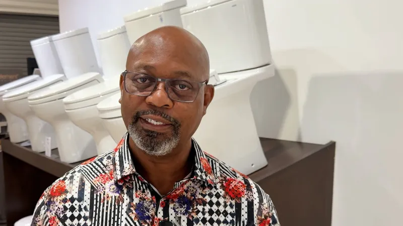 Man with glasses in a floral patterned shirt stands in front of a row of white toilets in a showroom.