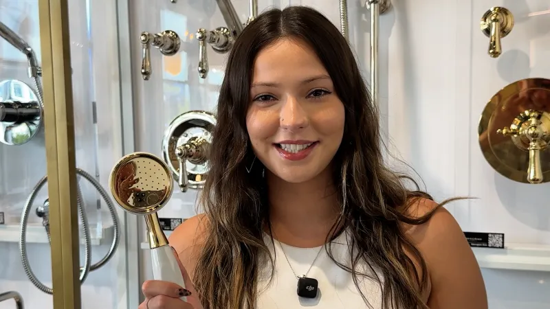 Smiling woman holding a gold showerhead in a showroom with various brass and gold shower fixtures behind her.