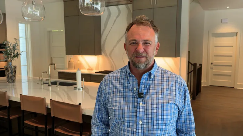 Man in blue checkered shirt standing in a modern kitchen with marble countertop and brown chairs.