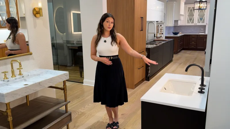 Woman in black skirt and white top presenting modern kitchen with marble sink, wood cabinets, and hardwood floors.