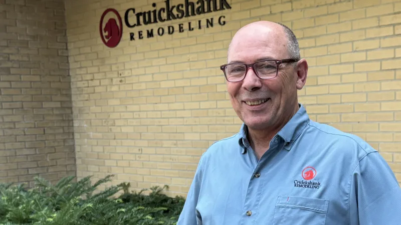Smiling man in blue Cruickshank Remodeling shirt stands beside yellow brick building with company logo.