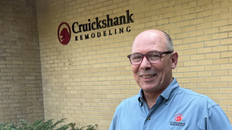 Smiling man in blue Cruickshank Remodeling shirt stands beside yellow brick building with company logo.