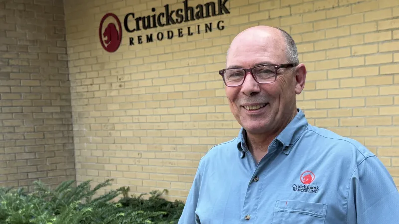 Smiling man in blue Cruickshank Remodeling shirt stands beside yellow brick building with company logo.