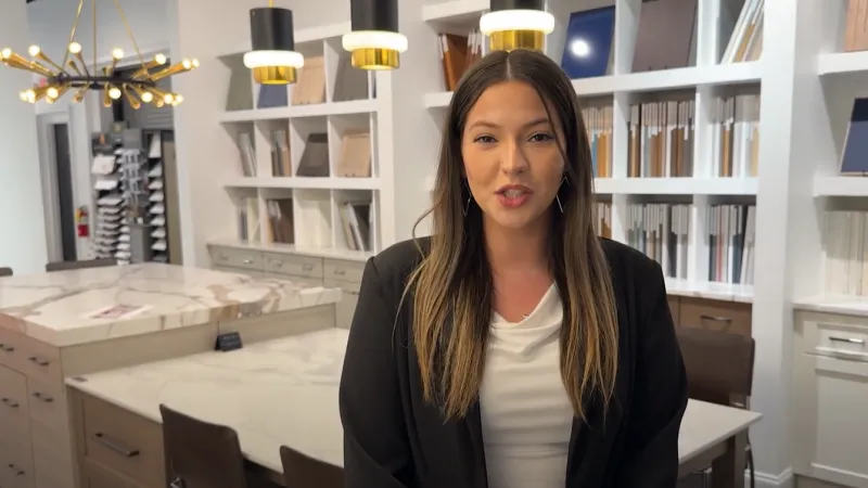 Woman in a black blazer standing in a modern showroom with marble countertops and samples on shelves behind her.