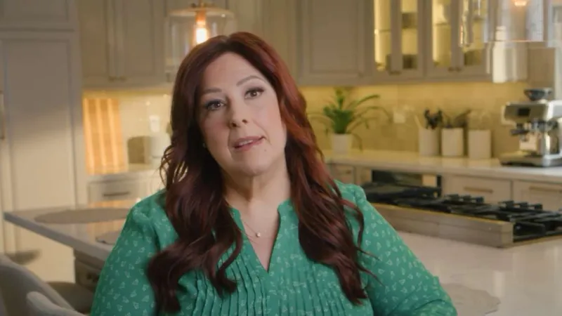 Woman with long red hair and green blouse sitting in a modern kitchen with marble countertops and gas stove.
