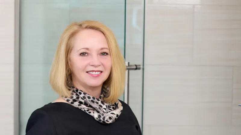 Smiling blonde woman wearing black top and patterned scarf standing in modern tiled room.