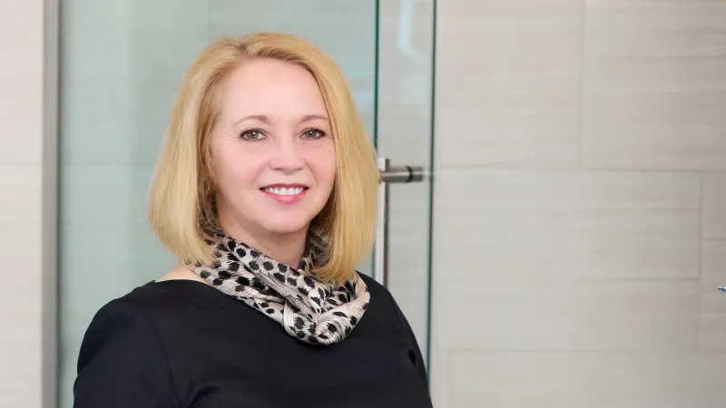 Smiling blonde woman wearing black top and patterned scarf standing in modern tiled room.