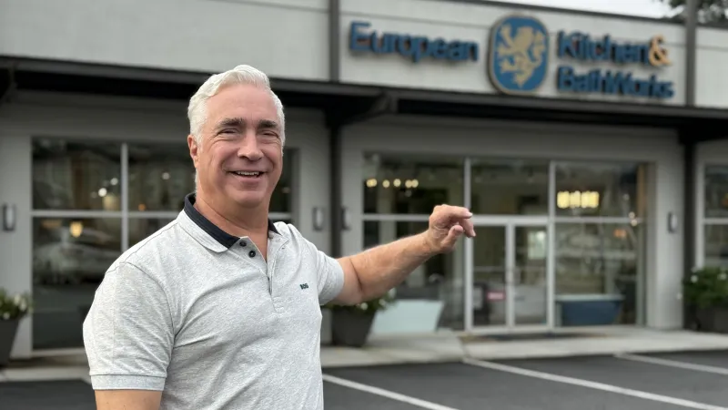 Smiling man in gray polo shirt pointing at European Kitchen & BathWorks storefront with glass windows
