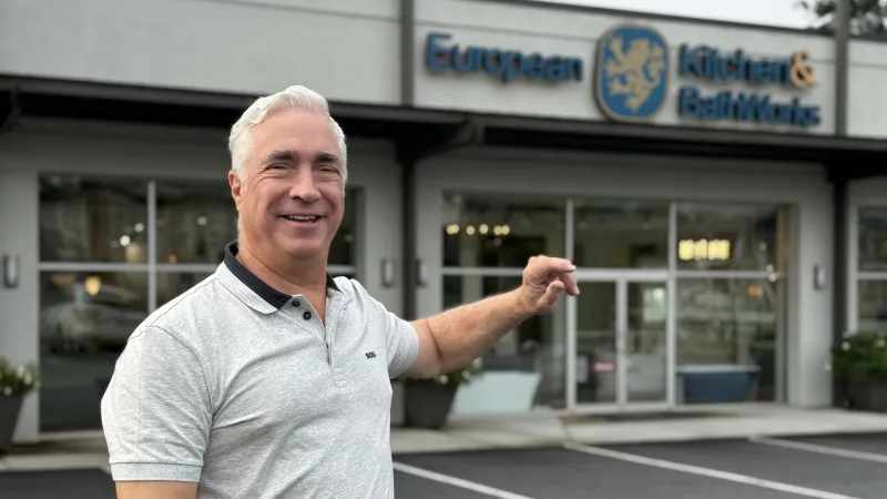 Smiling man in gray polo shirt pointing at European Kitchen & BathWorks storefront with glass windows
