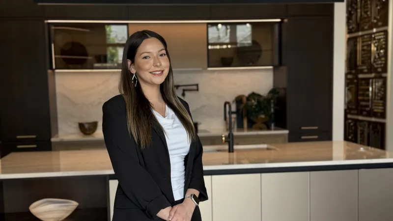 Smiling woman in black blazer and white shirt standing in a modern kitchen with marble counter and dark cabinets