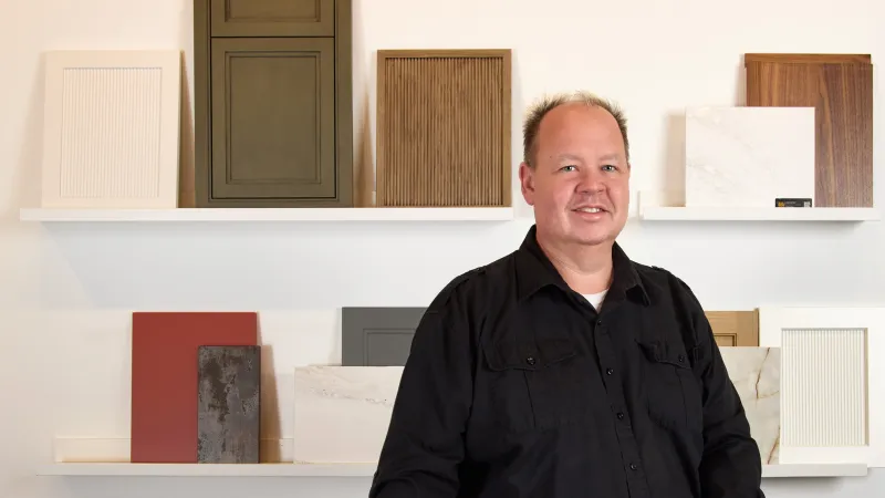 Man in black shirt standing in front of shelves displaying various wood and stone sample panels indoors.