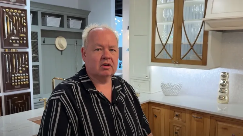 Man in a black striped shirt speaking in a modern kitchen with wooden cabinets and glass shelves.