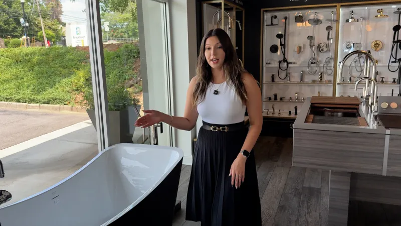 Woman presenting a modern freestanding bathtub in a showroom with various bathroom fixtures and natural light.