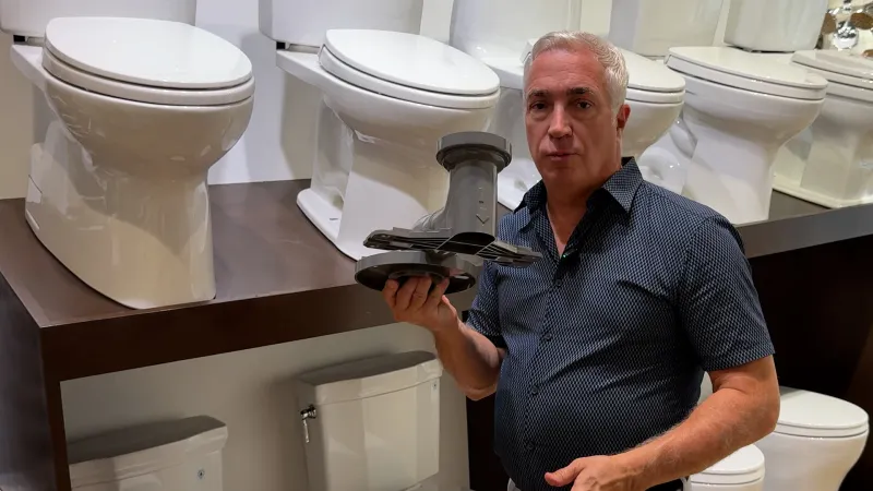 Man displaying a toilet flush valve in a showroom with multiple white toilets on shelves behind him