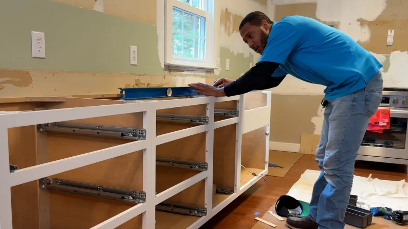 Man installing and leveling white kitchen cabinet frames in a partially renovated kitchen space.