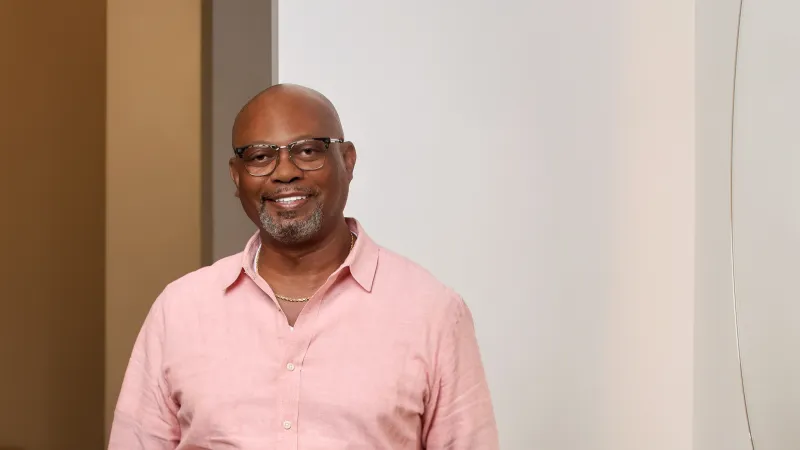 Smiling bald man wearing glasses and a pink shirt standing indoors against a neutral background.
