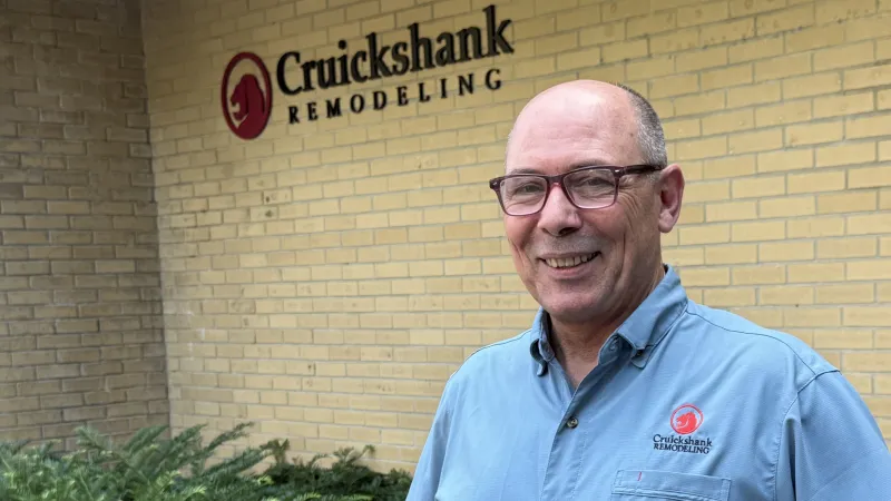 Smiling man in blue shirt stands outside building with Cruickshank Remodeling logo on brick wall and greenery nearby