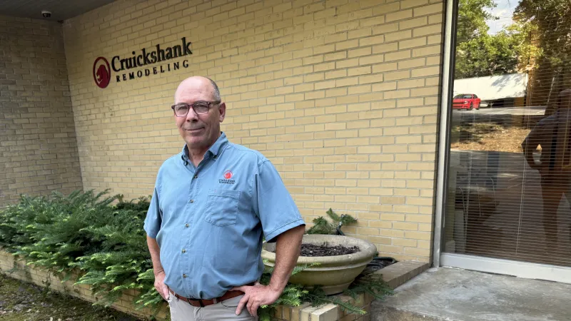 Man in blue shirt standing confidently in front of Cruickshank Remodeling office building with brick wall background.