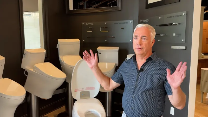 Man explaining various modern toilet models displayed in a showroom with dark brown walls and accessories panels.