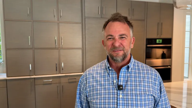 Smiling man in blue checkered shirt standing in modern kitchen with gray cabinets and built-in oven.