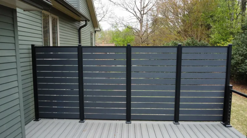 Modern black horizontal slat privacy fence on a gray deck attached to a green house exterior