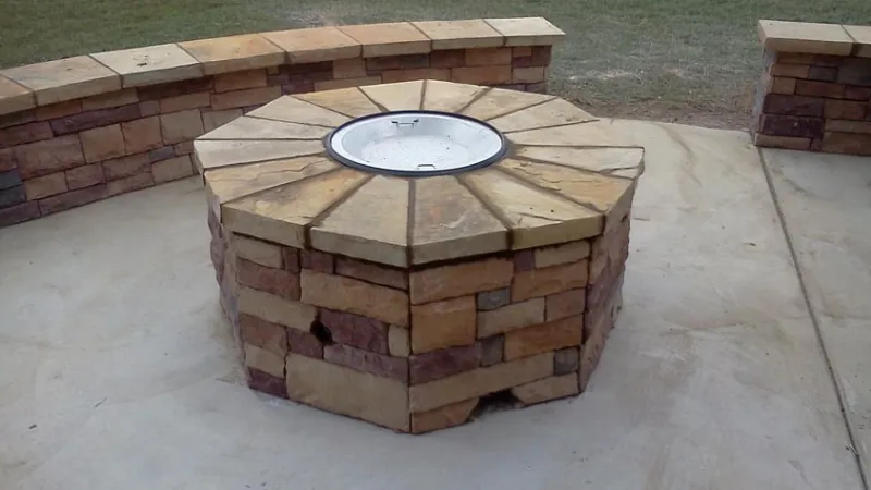 Stone outdoor fire pit with curved stone seating on concrete patio in a grassy backyard.