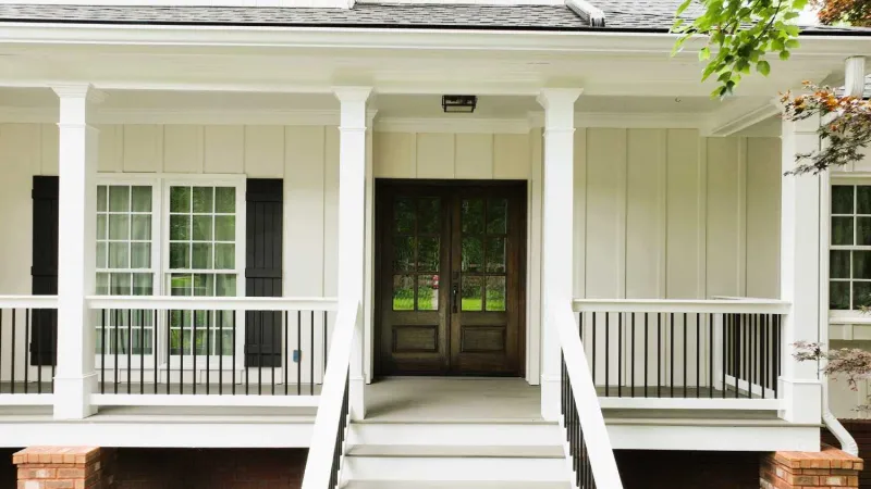 Front porch of a white house with wooden double doors, black railings, and brick foundation.
