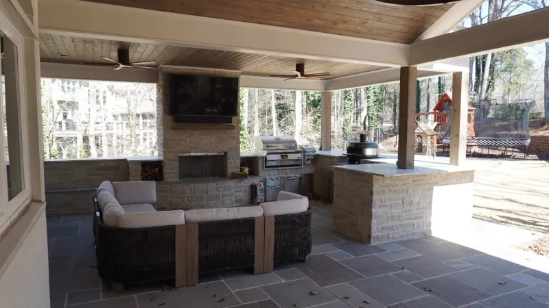 Covered outdoor patio with stone fireplace, built-in grill, L-shaped sofa, ceiling fans, and a play area visible outside.