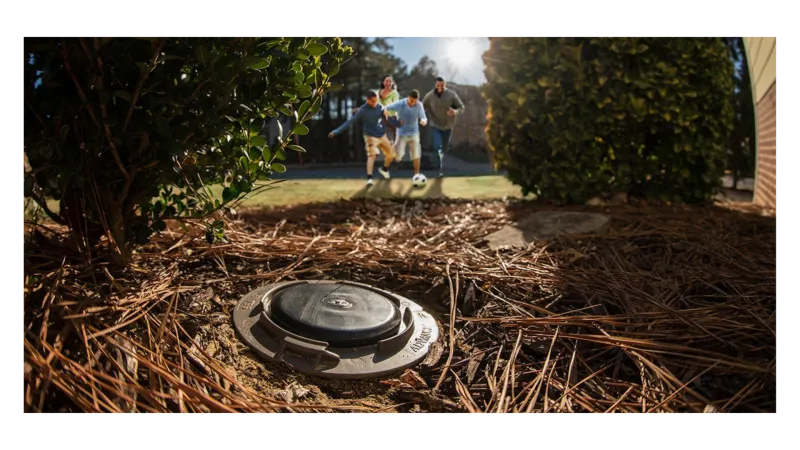 Backyard irrigation valve with family playing soccer in sunlit garden surrounded by bushes and pine needles.