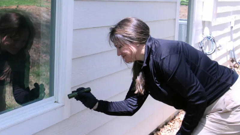 Woman inspecting window exterior with flashlight wearing black gloves and jacket during daytime outside house